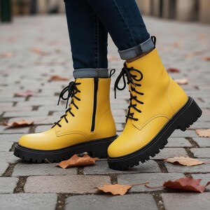 May include: Bright yellow ankle boots with black laces and chunky black soles. The boots feature a side zip and are worn with blue jeans. The boots are on a brick surface with fallen leaves.