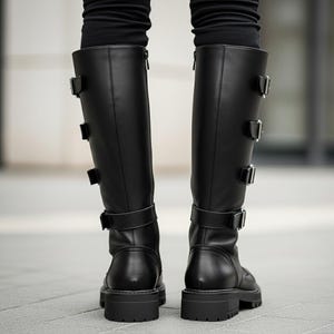 以下が含まれることがあります： 複数のバックルストラップと分厚いソールが付いた黒のニーハイブーツ。 ブーツはサイドにジッパーが付いており、滑らかな革のような素材で作られています。 後ろから見たブーツ。