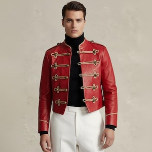Könnte beinhalten: Rote Lederjacke mit goldfarbenen Knöpfen und Besatz. Die Jacke hat einen hohen Kragen und ein militärisch inspiriertes Design. Das Model trägt einen schwarzen Rollkragenpullover und eine weiße Hose.