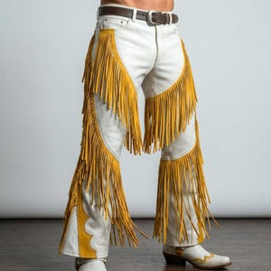 Puede incluir: Chaps de cuero blanco con largos flecos de color amarillo mostaza a lo largo de las piernas. La prenda incluye un cintur&oacute;n de cuero marr&oacute;n oscuro con una hebilla plateada en la cintura y se lleva sobre pantalones y botas de cuero blanco.