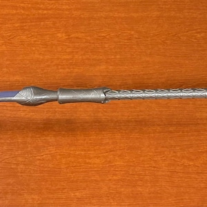 May include: A silver-colored staff with intricate detailing, featuring a bulbous end with a purple center and a similar design on the opposite end. The staff is supported by two small, dark gray stands.