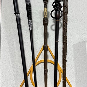 May include: A collection of five wands displayed on a wall-mounted holder. The wands vary in color and design, with details like carved handles and decorative elements. The holder is a white shelf with a gold triangle.