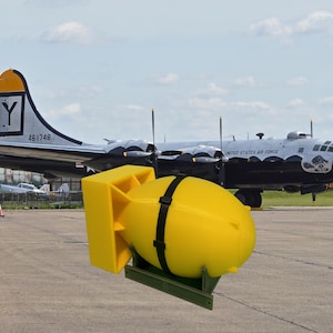 Op de afbeelding: Een gele bomreplica met zwarte banden, gemonteerd op een groene standaard, voor een vintage bommenwerper van de US Air Force. Het vliegtuig is zilver en zwart, met de tekst "UNITED STATES AIR FORCE" op de zijkant.