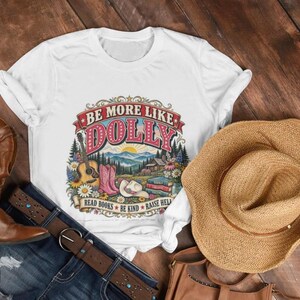 May include: A white t-shirt featuring a colorful graphic with the text BE MORE LIKE DOLLY, READ BOOKS, BE KIND, RAISE HELL. The design includes a guitar, pink cowboy boots, a cowboy hat, books, flowers, and a mountain landscape scene.