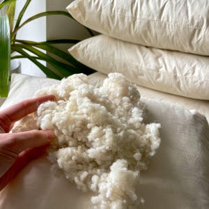 May include: Close-up of a pile of fluffy, white pillow filling next to a pillow. The filling is soft and cloud-like. Several pillows are stacked in the background, suggesting a bedding or home decor theme.