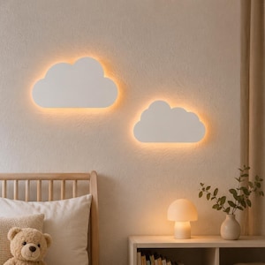 Könnte beinhalten: Zwei weiße, wolkenförmige Wandleuchten an einer Wand, die jeweils ein sanftes, warmes Licht nach hinten abgeben. Darunter steht eine pilzförmige Tischleuchte auf einem Regal neben einer Vase mit grünen Blättern und einem Teddybären.