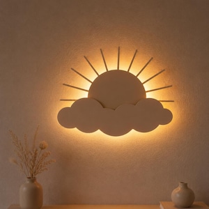 Könnte beinhalten: Eine Wandleuchte in Form einer Sonne, die hinter einer Wolke aufgeht. Die Leuchte hat eine matte, beige Oberfläche und strahlt ein warmes, leuchtendes Licht hinter der Silhouette aus, wodurch ein sanfter Lichtschein an der Wand entsteht.