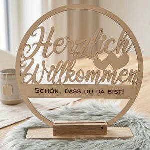 Könnte beinhalten: Ein Holz-Willkommensschild mit den Worten "Herzlich Willkommen" und "Schön, dass du da bist!" in einem Kreis ausgeschnitten. Das Schild enthält zwei Herzformen und steht auf einem Holzsockel.