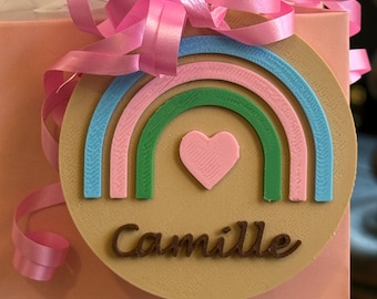 May include: A gift tag with a rainbow design in pastel colors, a pink heart, and the name "Camille" in brown script. The tag is attached to a pink gift bag with a pink ribbon bow.