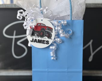 May include: A light blue gift bag with a matching twisted paper handle. A white tag with the name "JAMES" and a red monster truck graphic is attached with white curly ribbon. The bag is filled with white tissue paper.