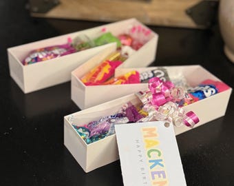May include: Three white rectangular gift boxes filled with colorful candies and wrapped with pink and white ribbons. A gift tag with the name "MacKenzie" and "Happy Birthday" is attached to one box.