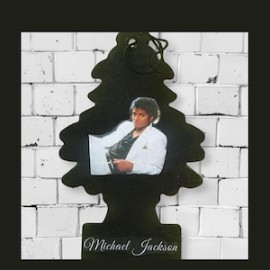 May include: Black air freshener in the shape of a Christmas tree with a photo of Michael Jackson. The photo shows Michael Jackson wearing a white suit. The name "Michael Jackson" is written in cursive at the bottom.