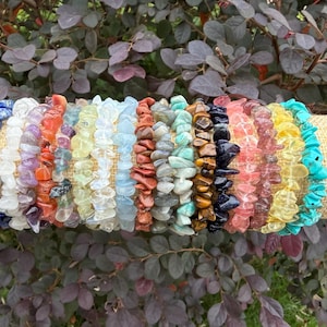 May include: A collection of colorful gemstone chip bracelets displayed on a tan fabric-covered cylinder. The bracelets feature a variety of colors, including blue, white, orange, green, brown, black, pink, yellow, and turquoise.