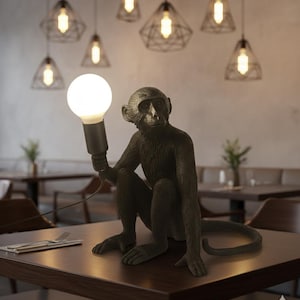 Puede incluir: Lámpara de mesa con forma de mono en bronce oscuro que sostiene una bombilla blanca redonda y grande. La lámpara está sobre una mesa de madera oscura en un restaurante. Encima, varias lámparas colgantes con bombillas expuestas cuelgan del techo.