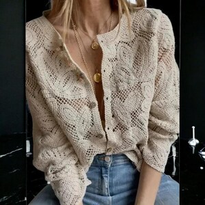 Puede incluir: Un cárdigan de crochet beige con botones y mangas largas. El diseño de tejido abierto revela un patrón floral. El cárdigan se combina con vaqueros azules y collares dorados.