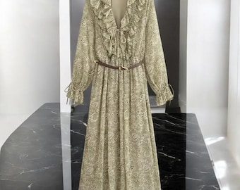 May include: A long-sleeved, olive green dress with a floral pattern. The dress features a V-neckline with ruffled trim, a tie closure, and a brown belt. The sleeves have ruffled cuffs. The dress is made of a lightweight fabric.