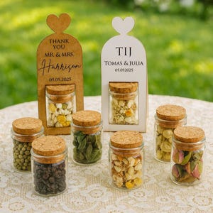 Op de afbeelding: Kleine glazen potjes met kurken deksels, gevuld met gedroogde bloemen en kruiden. Twee houten borden in de vorm van een hart met de tekst "Thank You Mr & Mrs Harrison 05.05.2025" en "TIJ Tomas & Julia 05.05.2025".