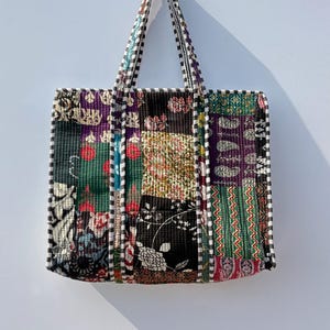 May include: A patchwork tote bag with a variety of floral and geometric patterns in shades of green, red, purple, and black. The bag has a black and white checkered trim and handles.