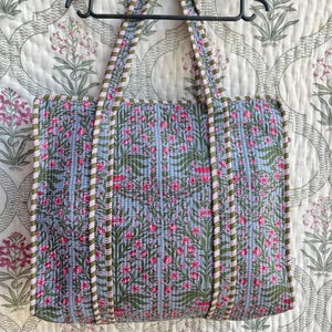 Peut inclure: Un sac fourre-tout matelassé avec un motif floral rose et vert sur fond bleu clair. Le sac a des poignées et des bordures assorties. Il est suspendu à un cintre noir, sur un fond à motifs floraux.