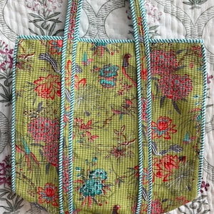 May include: A quilted tote bag with a floral pattern in shades of green, red, and teal. The bag has teal trim and handles with a white and teal rope design. The bag is hanging on a brown hanger.