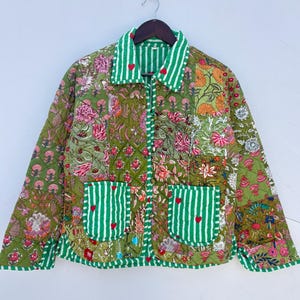 Puede incluir: Una chaqueta acolchada con un patchwork de patrones florales verdes y detalles de rayas blancas y verdes. La prenda incluye un cuello, mangas largas y dos bolsillos delanteros, todos decorados con pequeños detalles en forma de corazón rojo.
