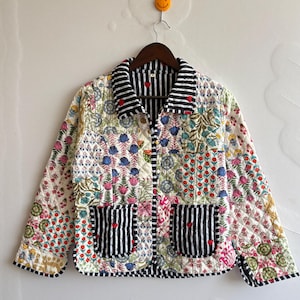 Op de afbeelding: Een gewatteerd jack met een patchwork van bloemmotieven in rood, blauw, groen en geel. Het kledingstuk heeft een zwart-wit gestreepte kraag, manchetten en twee voorzakken, elk geaccentueerd met een klein rood hartdetail.