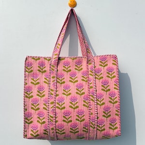 May include: Pink tote bag with a floral pattern. The bag features a repeating design of purple flowers with green stems on a pink background. The handles and edges are trimmed with a red and white striped fabric. The bag is hanging on a light blue wall.