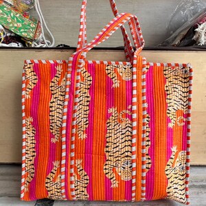 May include: A rectangular tote bag with a vibrant orange and pink striped pattern. The bag features a repeating tiger print and white vertical stripes. The handles and trim are striped in orange, pink, and white. The bag is made of a quilted fabric.