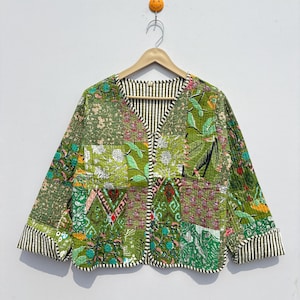 May include: A quilted jacket with a patchwork design in shades of green, featuring floral and geometric patterns. The jacket has a V-neck and striped cuffs and trim. The jacket is hanging on a wooden hanger.