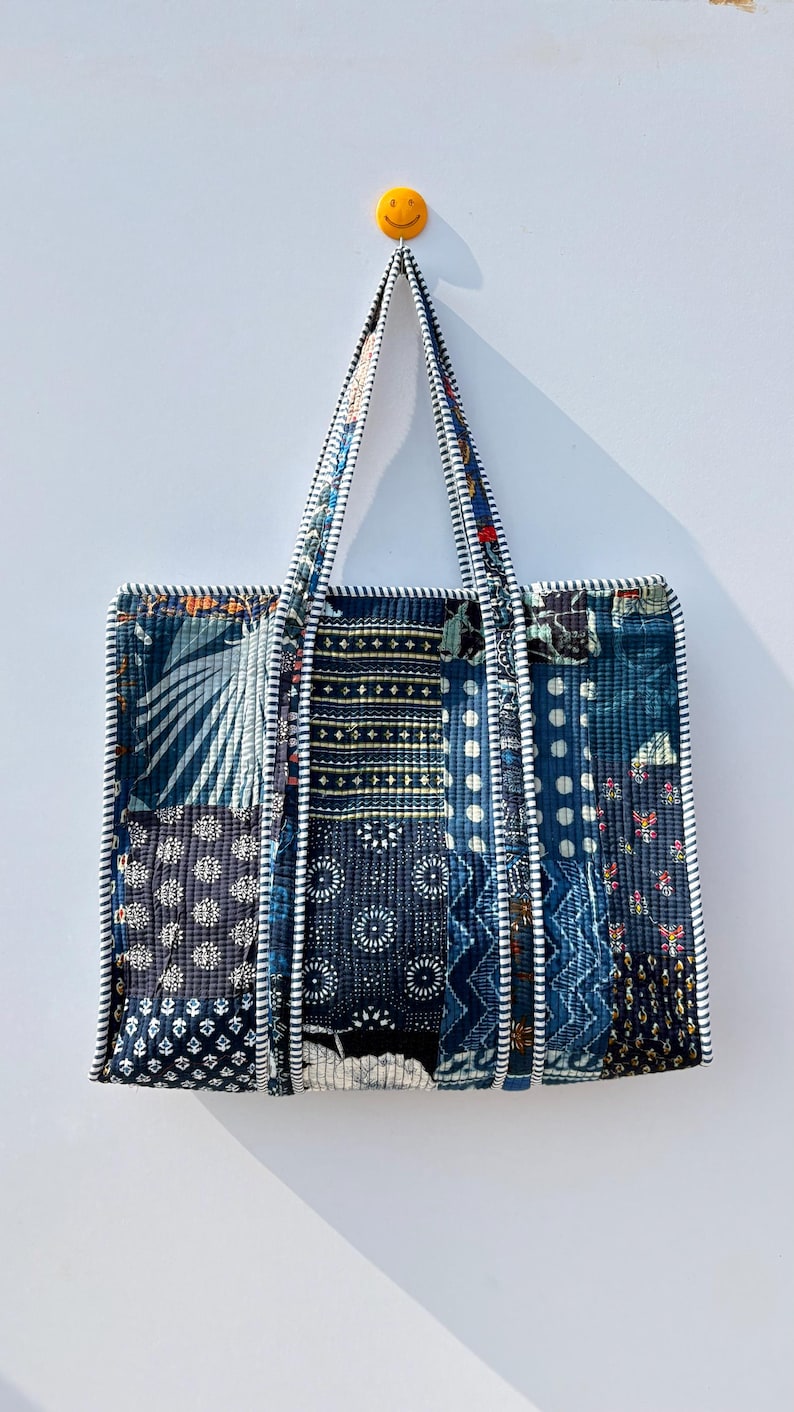 May include: A patchwork tote bag with a variety of blue fabric patterns. The bag has a rectangular shape with two handles and a white and black striped trim. A yellow smiley face is on the wall behind the bag.