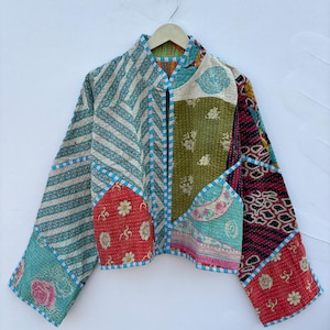 May include: A cropped jacket featuring a patchwork design of various fabrics in shades of red, green, blue, and white. The garment displays multiple patterns including floral prints, stripes, and geometric shapes, finished with blue and white striped trim.