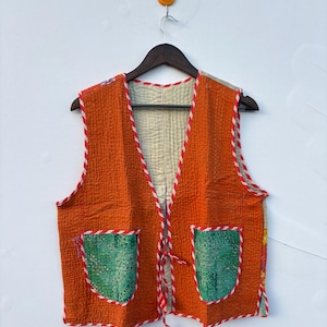 May include: A sleeveless waistcoat featuring a vibrant orange quilted fabric with a light cream interior lining. The garment includes two green patterned patch pockets and is finished with red and white striped trim along the edges and tie closures.