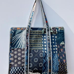 May include: A patchwork tote bag with a variety of blue and white patterned fabrics. The bag has two handles and is trimmed with white piping. The bag is hanging on a white wall.