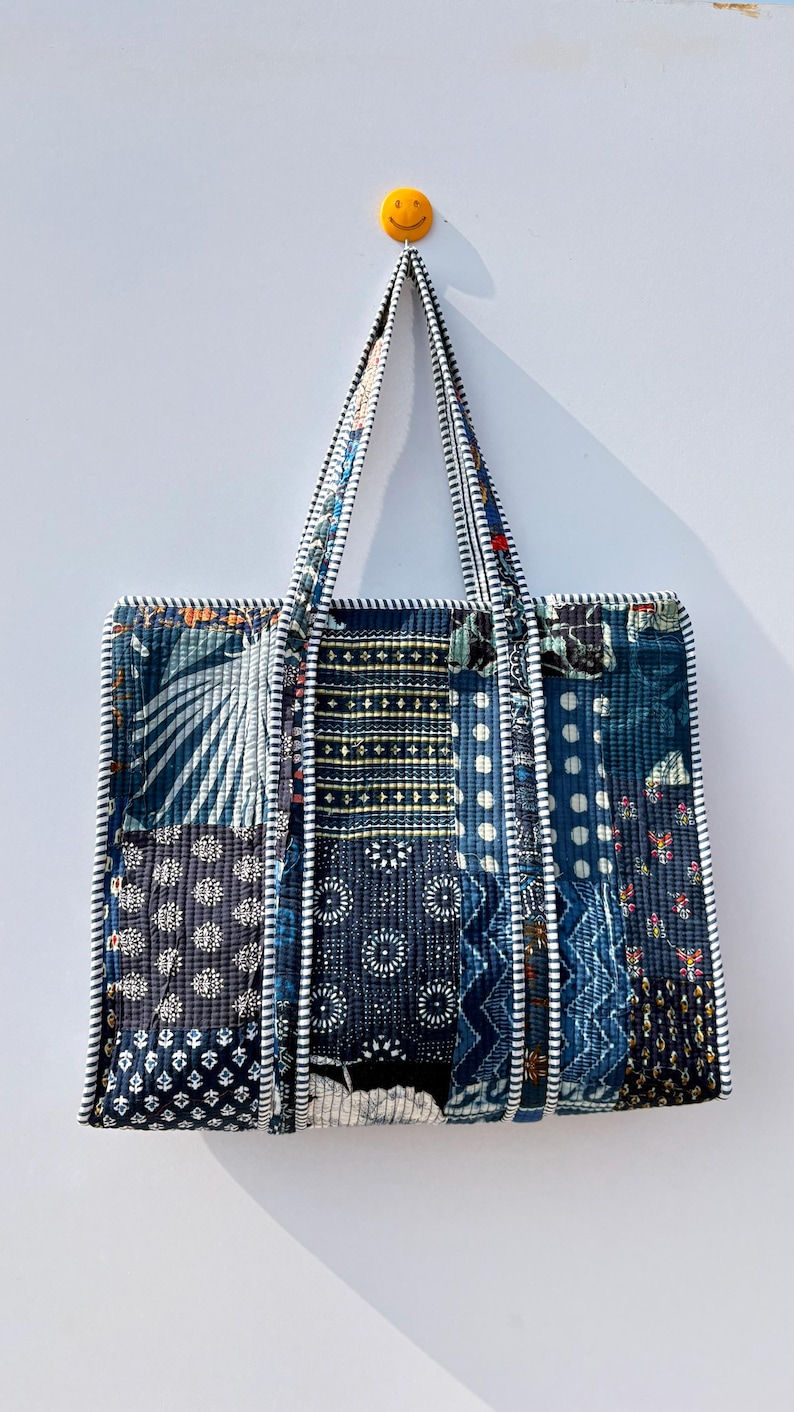 May include: A patchwork tote bag with a variety of blue and white patterned fabrics. The bag has two handles and is trimmed with white piping. The bag is hanging on a white wall.