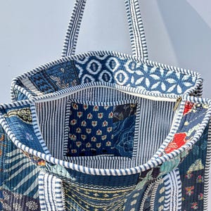 May include: Open tote bag with a patchwork design in shades of blue, white, and navy. The bag features various patterns, including floral and geometric shapes. The interior has a striped lining and a small pocket. The straps are white with blue stripes.