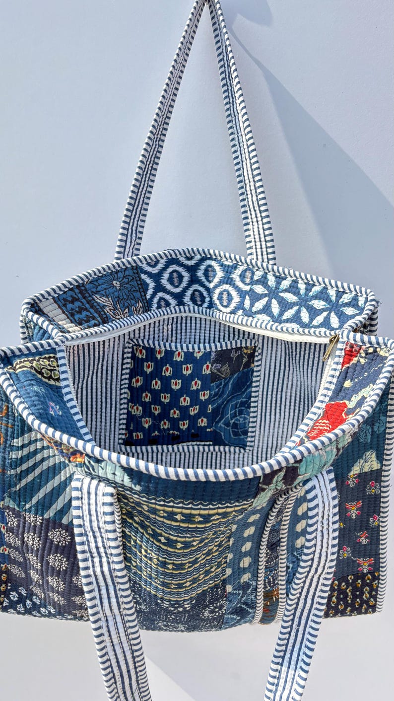 May include: Open tote bag with a patchwork design in shades of blue, white, and navy. The bag features various patterns, including floral and geometric shapes. The interior has a striped lining and a small pocket. The straps are white with blue stripes.
