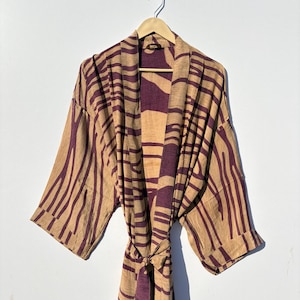 May include: A beige and purple patterned kimono robe hanging on a wooden hanger. The robe features a striped design in shades of beige and deep purple. The robe is tied at the waist with a matching belt.