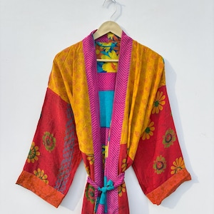 Op de afbeelding: Een kleurrijke, patroonjas met een striksluiting. De jas heeft een mix van rode, gele, roze en blauwe stoffen met bloemen- en geometrische patronen. De mouwen en de rand hebben een contrasterende oranje rand. De jas hangt aan een houten hanger.