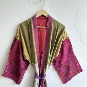 May include: A kimono-style robe featuring a patchwork design with olive green, magenta, and light gray panels. The garment has wide sleeves, a magenta patterned collar, and a matching tie belt at the waist, all displayed on a wooden hanger.