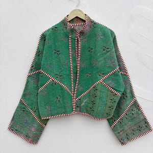 May include: A green quilted jacket featuring a mandarin collar and wide sleeves. The garment is constructed with visible patchwork seams outlined in a red and white striped trim, and it displays a subtle, repeating floral pattern throughout.