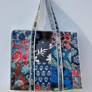 May include: A quilted tote bag with a patchwork design. The bag features various floral and geometric patterns in shades of blue, red, and white. The handles and edges are trimmed with a striped fabric.
