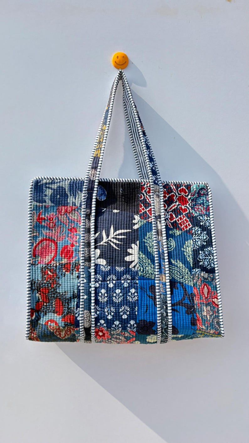 May include: A quilted tote bag with a patchwork design. The bag features various floral and geometric patterns in shades of blue, red, and white. The handles and edges are trimmed with a striped fabric.