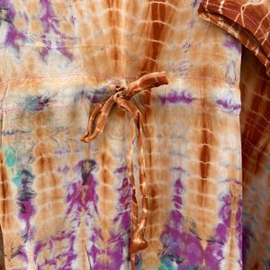 Puede incluir: Prenda te&ntilde;ida con tinte anudado en tonos naranja, marr&oacute;n, morado y turquesa. La tela presenta una cintura con cord&oacute;n y un nudo. El dise&ntilde;o incluye patrones verticales y florales, creando una est&eacute;tica bohemia. La textura de la prenda parece suave y fluida.