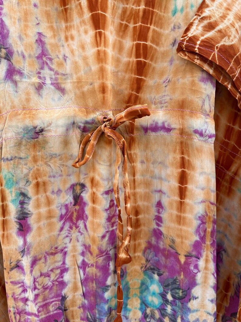 Puede incluir: Prenda te&ntilde;ida con tinte anudado en tonos naranja, marr&oacute;n, morado y turquesa. La tela presenta una cintura con cord&oacute;n y un nudo. El dise&ntilde;o incluye patrones verticales y florales, creando una est&eacute;tica bohemia. La textura de la prenda parece suave y fluida.