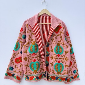 May include: A pink embroidered jacket with a reversible design. The outer layer features floral and pomegranate patterns in teal, red, and brown. The inside lining has red and white vertical stripes. The jacket is hanging on a wooden hanger.
