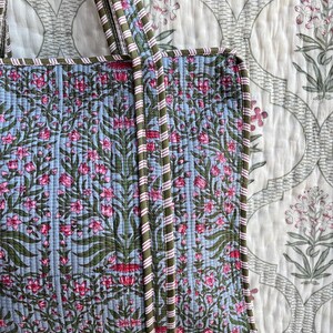 May include: A quilted tote bag with a floral pattern in pink and green on a light blue background. The bag has green and pink striped handles and trim. The background is a white floral patterned fabric.