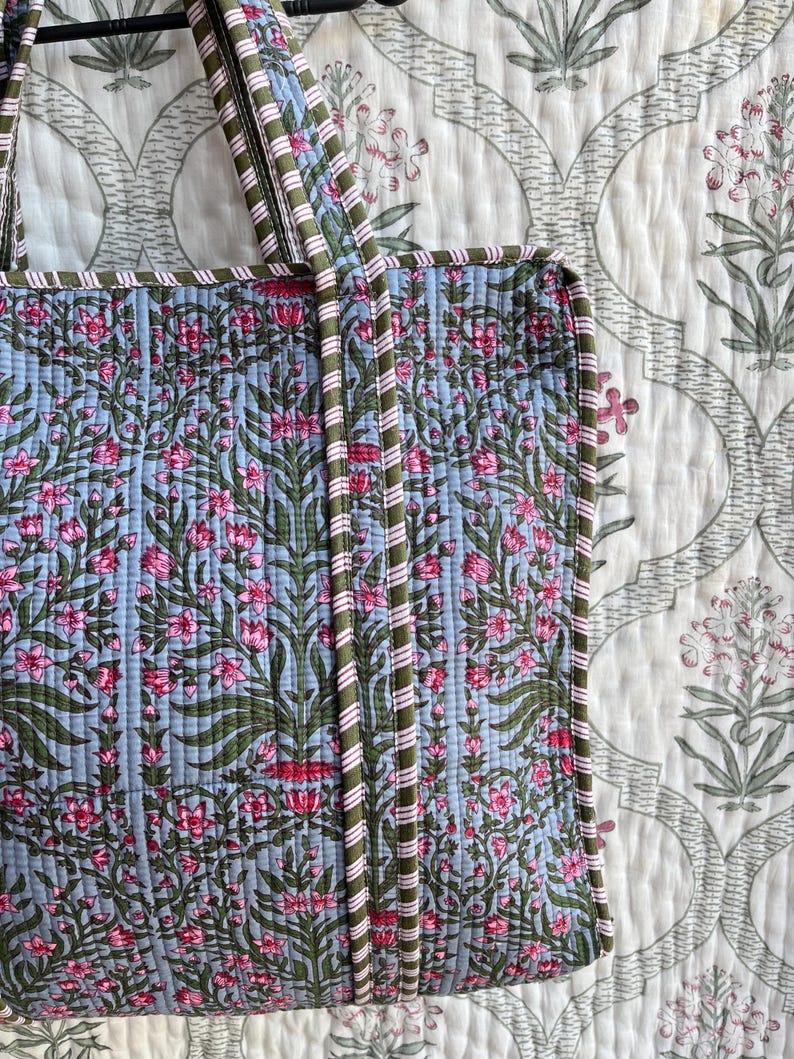 May include: A quilted tote bag with a floral pattern in pink and green on a light blue background. The bag has green and pink striped handles and trim. The background is a white floral patterned fabric.