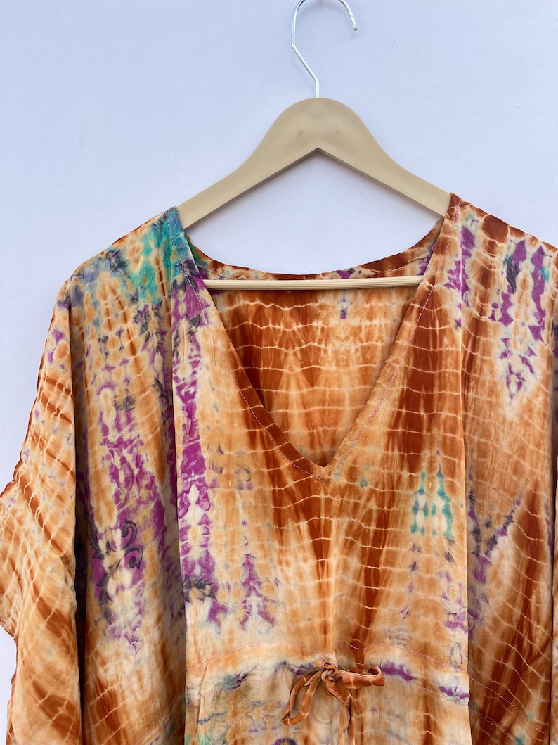 Puede incluir: Un caft&aacute;n te&ntilde;ido anudado en tonos naranja, marr&oacute;n, morado y turquesa cuelga de una percha de madera. La prenda con cuello en V presenta una cintura con cord&oacute;n y un patr&oacute;n colorido &uacute;nico. La tela parece ligera y fluida.