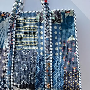 May include: A quilted tote bag with a patchwork design in shades of blue, white, and touches of red. The bag features various geometric and floral patterns. The handles and edges are trimmed with a striped fabric.