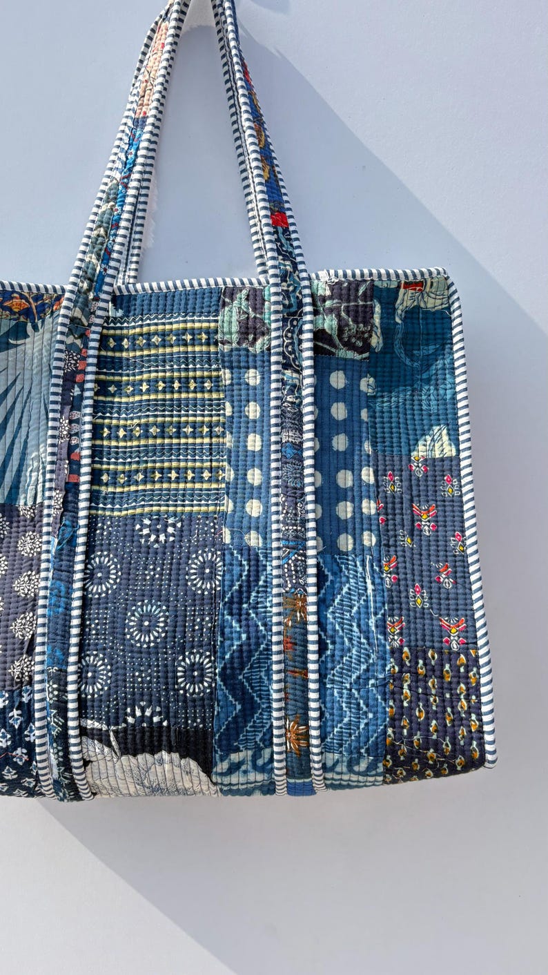 May include: A quilted tote bag with a patchwork design in shades of blue, white, and touches of red. The bag features various geometric and floral patterns. The handles and edges are trimmed with a striped fabric.